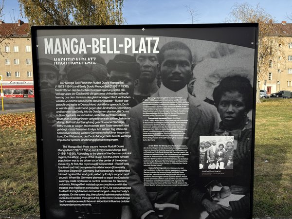 Schwarz-weiße Informationstafel mit dem Titel 'MANGA-BELL-PLATZ' und Text über Emily und Rudolf Douala Manga Bell sowie zwei historischen Fotos.