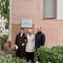 Drei Personen stehen nebeneinander vor einer Hauswand mit Gedenktafel und geschlossenen Rollläden, umgeben von Büschen.