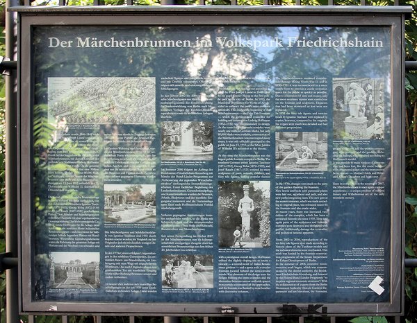 Informationstafel mit Text und historischen Fotos zum Märchenbrunnen im Volkspark Friedrichshain, teilweise von Baumblättern und Lichtreflexen überlagert