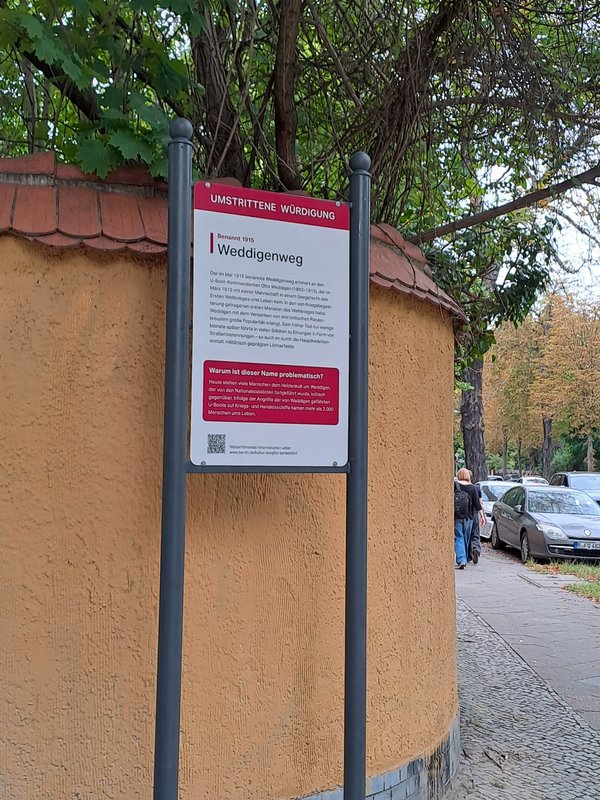 Informationsschild mit der Überschrift 'Umstrittene Würdigung' und dem Namen 'Weddigenweg' an einer gelben Mauer neben einem Gehweg mit parkenden Autos und Passanten
