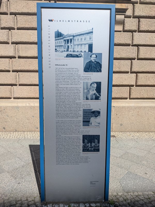 Informationstafel mit Text und historischen Schwarz-weiß-Fotografien vor einer Sandsteinfassade