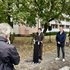 Eine Person im langen Mantel spricht an einem Rednerpult im Freien vor mehreren Zuhörenden unter Bäumen