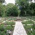 Blick auf einen symmetrisch angelegten Rosengarten mit gepflastertem Mittelweg und mehreren Bänken, umgeben von hohen Bäumen