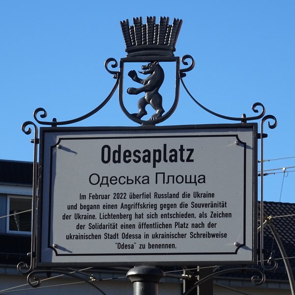 Metallschild mit Text zur Benennung des Platzes nach der ukrainischen Stadt Odessa, darüber das Berliner Wappen.