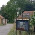 Straße mit altem Gebäude links, Laterne und Informationsschild am Wegesrand, umgeben von Bäumen und Sträuchern