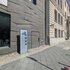 Straßenszene mit zwei nebeneinanderliegenden Gebäuden, eines mit glatter, modernes Fassadenmuster, das andere mit klassischer Steinfassade, davor ein Geschichtstafel auf dem Bürgersteig