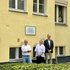 Drei Personen stehen vor einer gelben Hauswand mit einer Gedenktafel und mehreren Fenstern