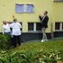 Drei Personen ziehen an Schnüren, um ein verhülltes Schild an einer gelben Hauswand zu enthüllen
