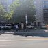 Straßenszene mit großem Baum, Informationstafel, mehreren geparkten Autos, einem Fahrrad an einem Schildpfosten und zwei Fußgängern auf dem Gehweg vor einem mehrstöckigen Wohngebäude