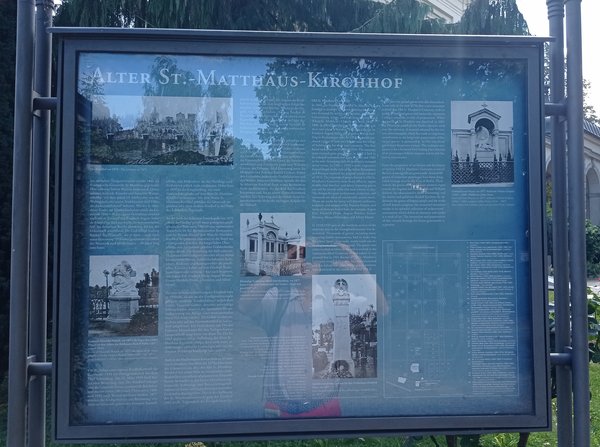 Informationstafel mit Text und historischen Fotos zum Alten St. Matthäus-Kirchhof in einem Metallrahmen im Freien