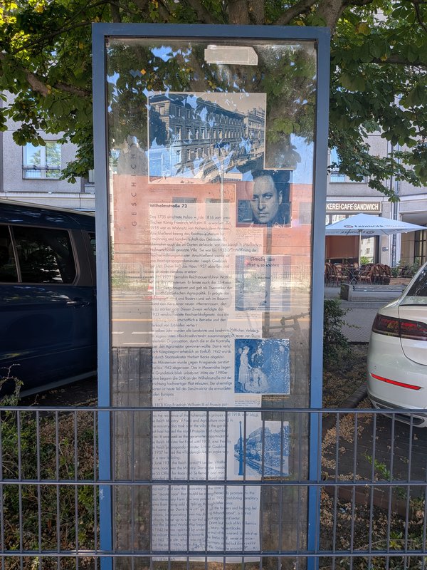 Informationsschild mit Text und historischen Fotos zu Wilhelmstraße 72 hinter Glas in einem blauen Rahmen an einem Straßenrand mit parkenden Autos und Bäumen im Hintergrund