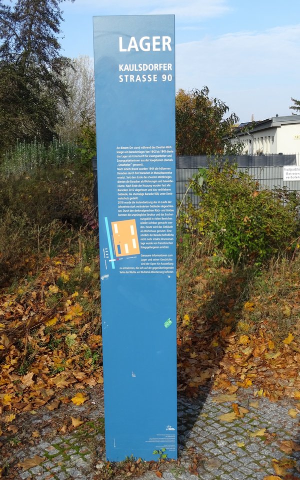 Blaue Stele mit Text und einer Grafik zum Lager Kaulsdorfer Straße, umgeben von Laub und Büschen.