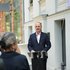 Staatssekretär Alexander Straßmeir in dunklem Sakko spricht an einem Rednerpult vor einem Gebäude mit Blumenkästen an den Fenstern