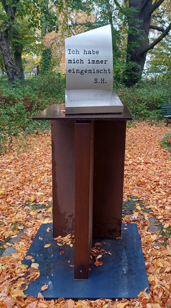 Stele mit der Inschrift 'Ich habe mich immer eingemischt S.H.' auf einem rostbraunen Metallgestell in einem herbstlichen Park mit Laub auf dem Boden