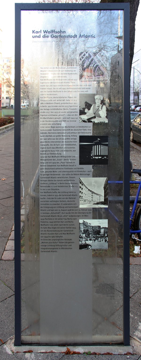 Eine Stele aus Acrylglas mit Text und Fotos zu Karl Wolffsohn, im Hintergrund sind Wohnhäuser, Gehweg und Autos.