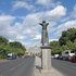 Statue einer stehenden Person mit erhobenen Armen auf einem Sockel an einer breiten Straße mit dem Brandenburger Tor im Hintergrund