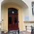 Eingangstür mit zwei dunkelroten Flügeln und der Hausnummer '21' über dem Bogen, umgeben von einem hellgelben Rahmen und einer hellen Fassade mit einem Informationsschild rechts