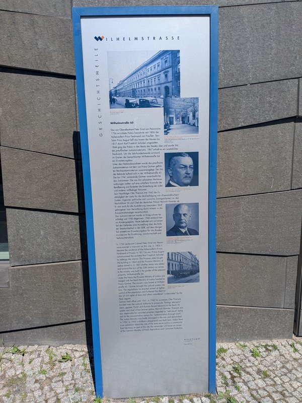 Geschichtstafel mit Text und mehreren historischen Fotos zu Wilhelmstraße 65 vor einer modernen Gebäudefassade