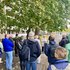 Mehrere Menschen stehen auf einem Gehweg unter Bäumen und hören einer Person an einem Rednerpult zu.