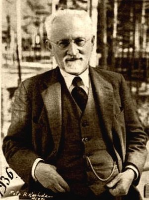 Portraitfoto von Simon Dubnow, sepia aus dem Jahr 1936. Er sitzt auf einem Stuhl und schaut direkt in die Kamera, trägt einen dunklen Anzug mit Krawatte.