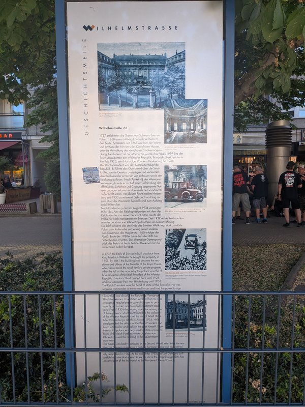 Informationstafel mit historischen Fotos und Text zur Wilhelmstraße in einem öffentlichen Bereich mit Menschen im Hintergrund
