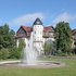 Brunnen mit Fontäne vor historischen Gebäuden und Bäumen auf einer Grünfläche unter blauem Himmel