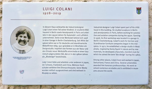 Weiße Gedenktafel mit Text zu Luigi Colani, seinem Leben und Werk, oben links sein Name und Lebensdaten, oben rechts ein Wappen