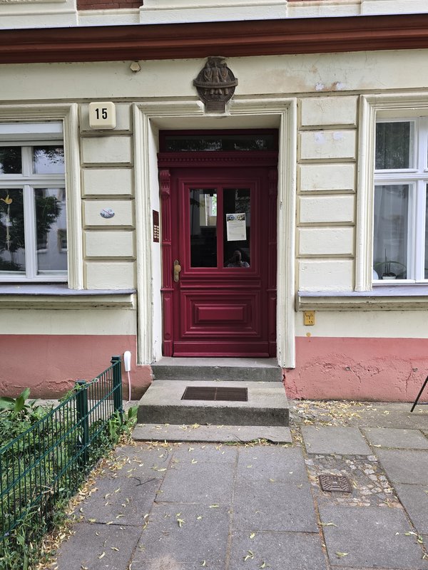 Eingangstür eines Altbaus mit roter Holztür, zwei Fenstern und Hausnummer 15, über der Tür eine Gedenktafel